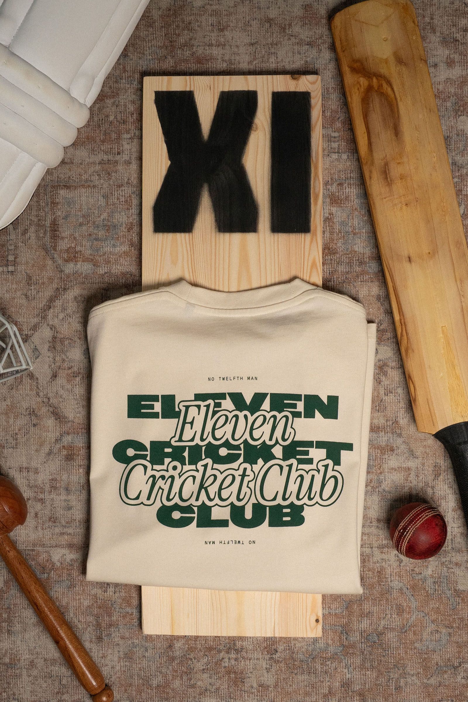 Cream Eleven Cricket Club T-shirt laid over an XI-branded cricket bat beside a red ball.