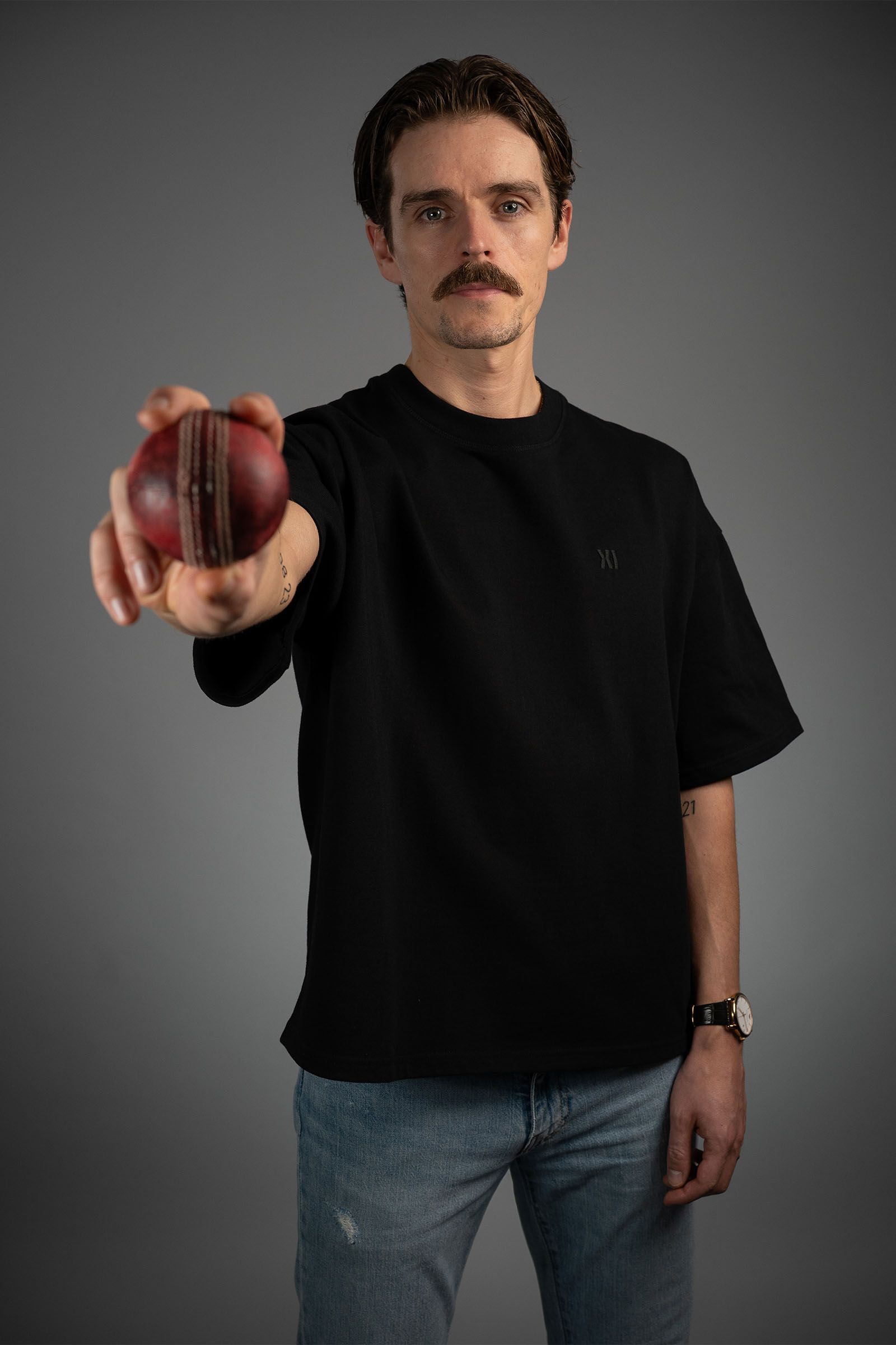 A model wearing a black Eleven Cricket Club T-shirt and holding a red cricket ball towards the camera.