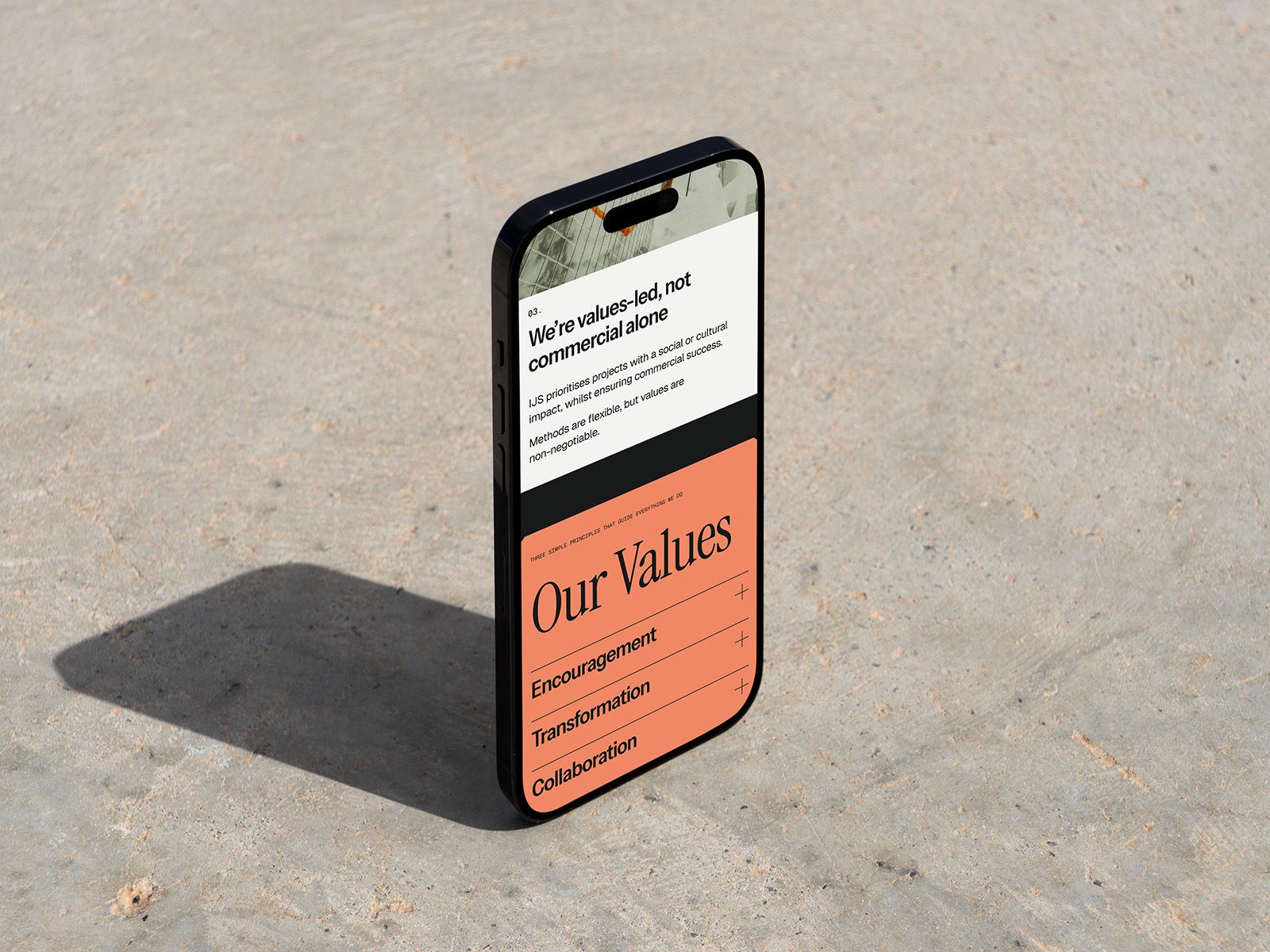 A phone standing on a sunlit concrete surface, displaying an Our Values section in black and coral.
