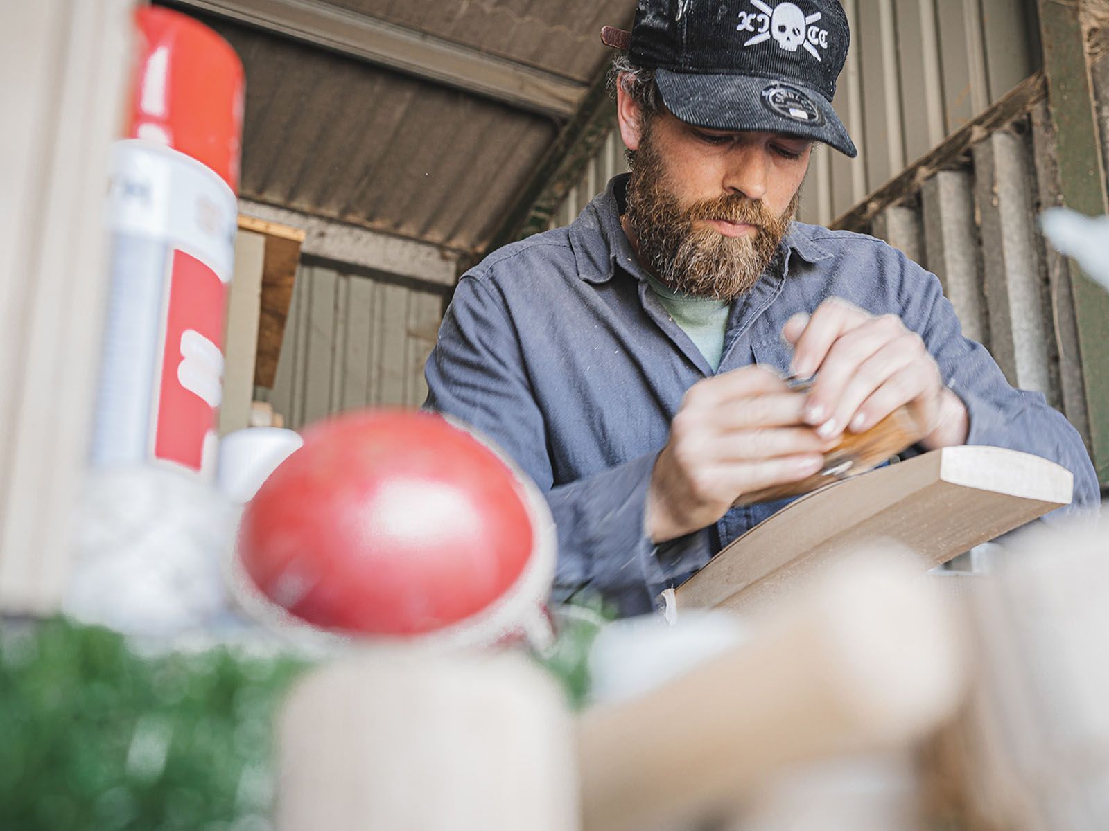 A bat maker working on a cricket bat at a bench, with a red cricket ball in the foreground.