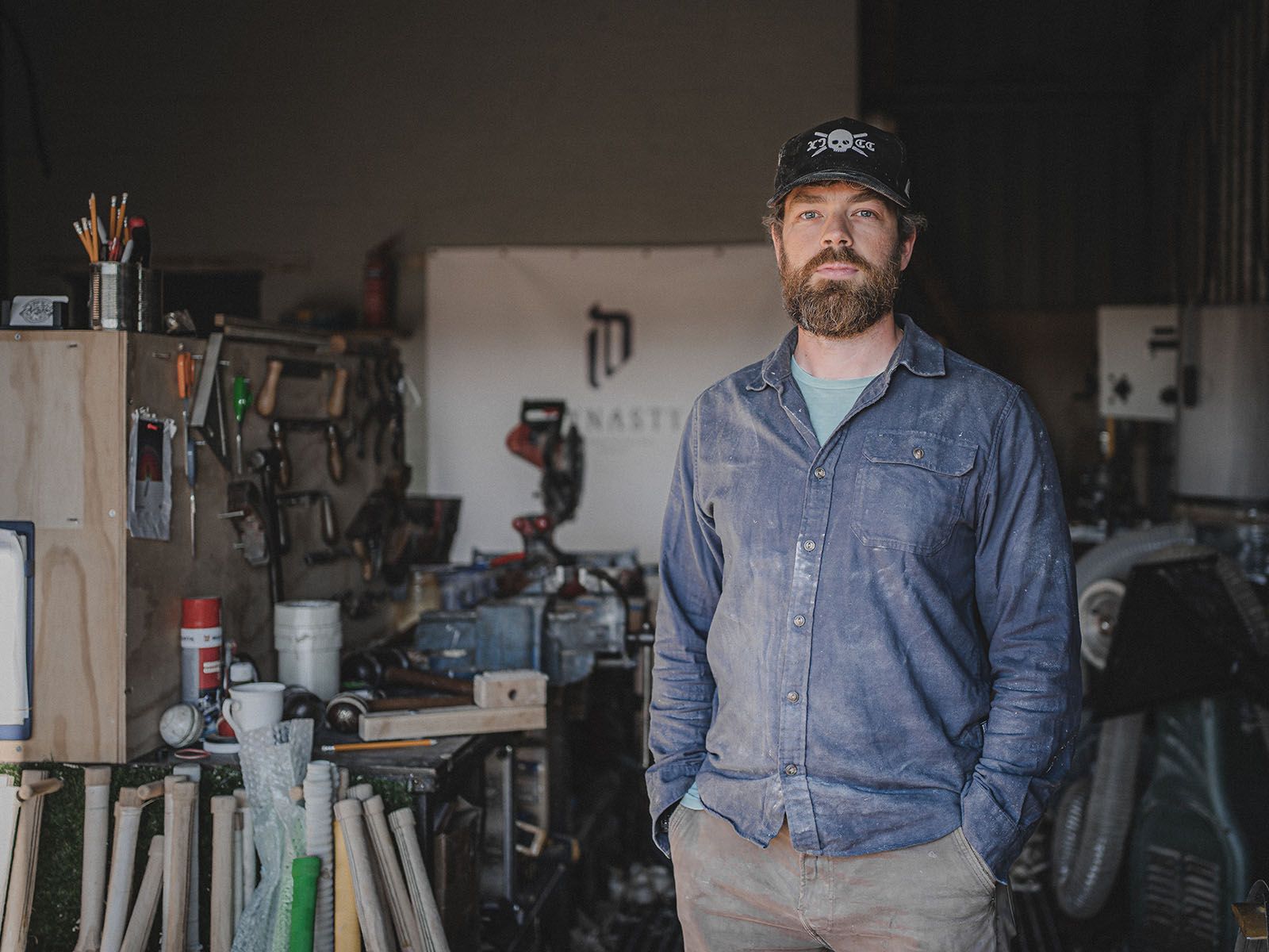 A cricket bat maker standing in a workshop wearing a blue work shirt and black cap.