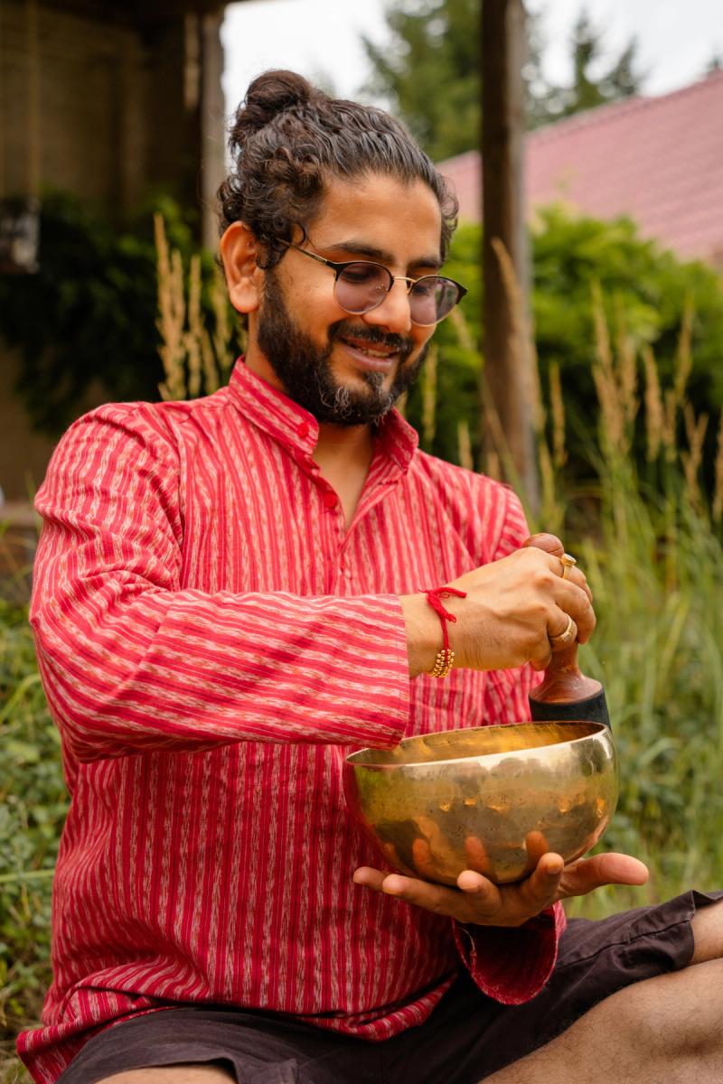 Dhiraj lächelt beim Spielen einer tibetischen Klangschale im grünen Garten, in rotem Kurta