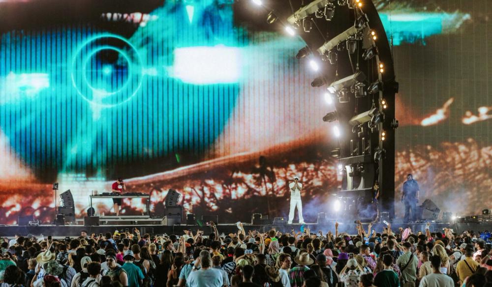 Skepta stands on stage in an all white outfit. There is a large screen behind him with an eye with a blue HUD overlay on it and a big crowd in front of the stage watching him perform.