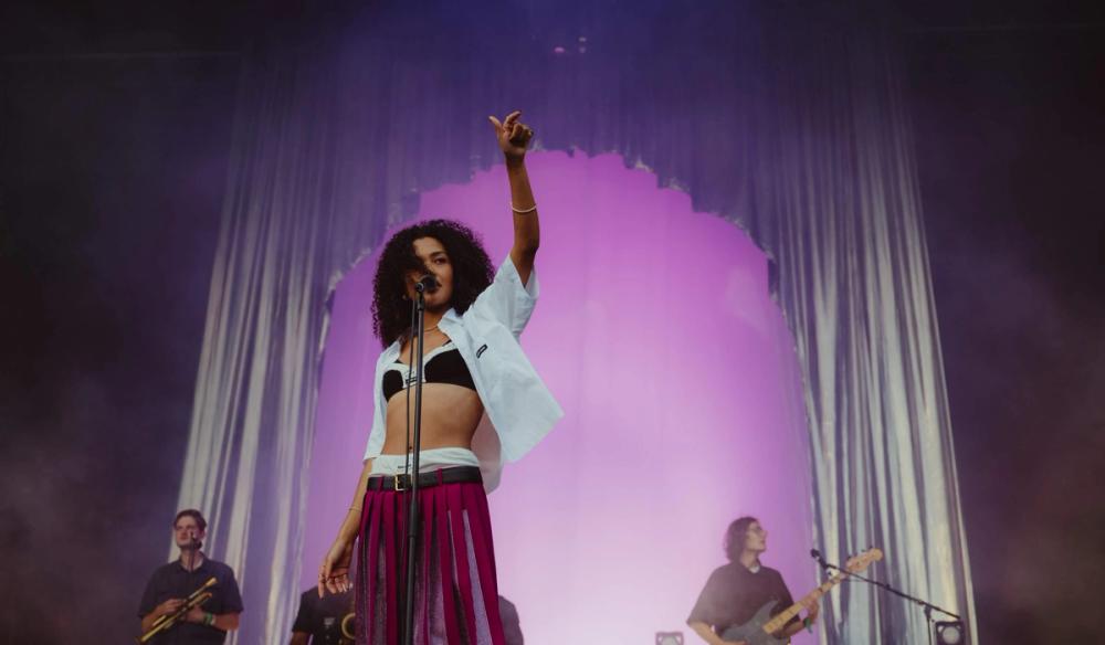 Olivia Dean stands on the left-hand side of the image with her arm raised, singing. Behind her is a metallic fabric arch with purple lighting inside the arch. Olivia wears a black bra, open white button down shirt, and maroon skirt.