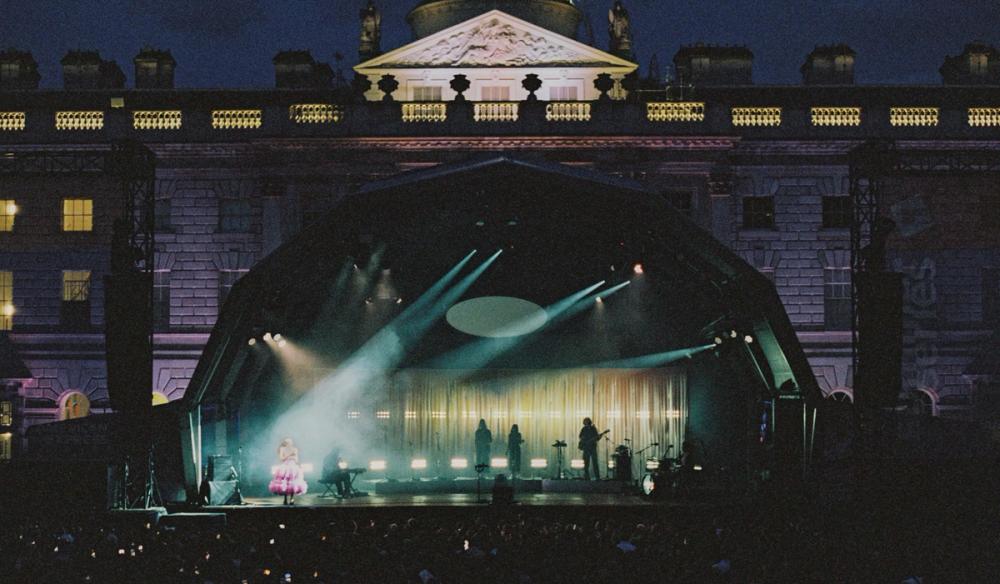 A half-dome shaped stage is shown in front of Somerset House at night. On the stage Olivia Dean is singing off-center to the left. There is a sheer curtain lit yellow along with four bandmates.
