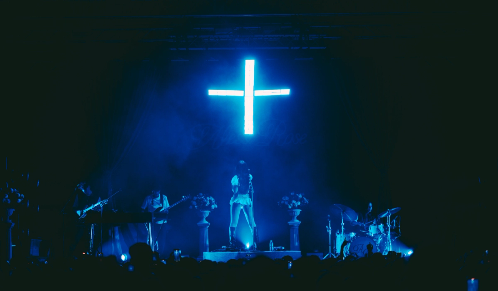 Alessi Stands with her back to the audience in blue lighting. A large glowing cross hangs above her with her bandmates playing on either side.