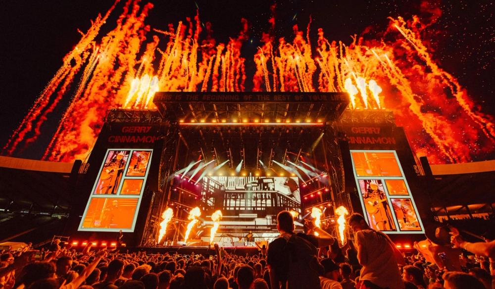 A wide image of a stage with comic book style graphics on the screen and pyro shooting off in the background