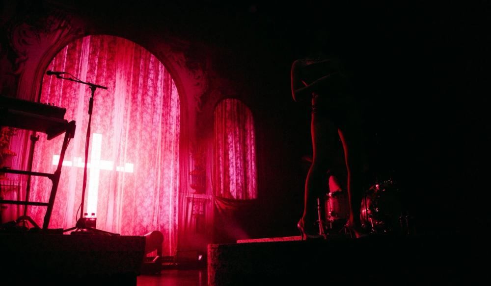 a pink cross illuminates lace curtains behind pop star Alessi Rose