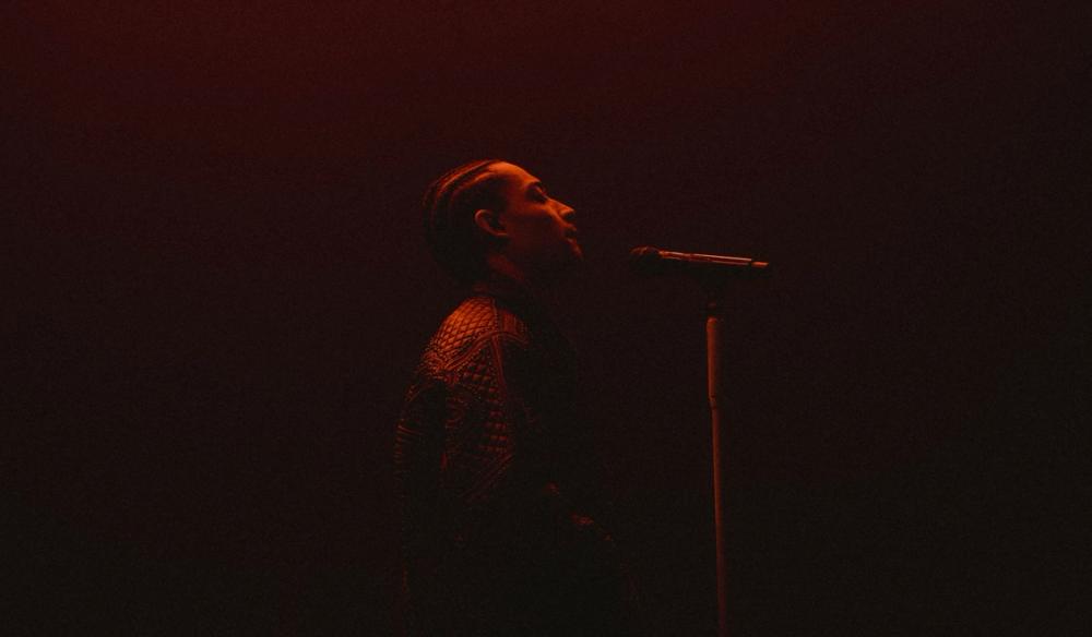 Loyle Carner stands sideways to camera and sings into a microphone in dark red lighting.