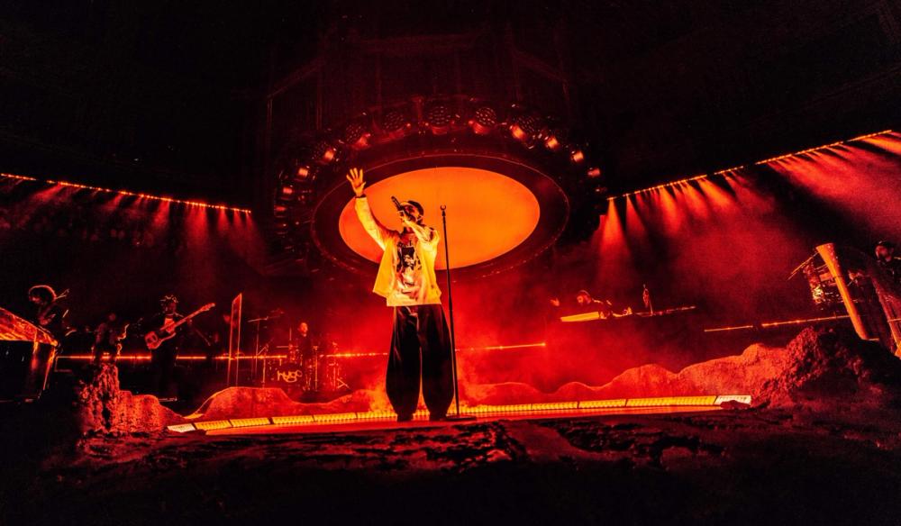 Artist Loyle Carner raps with arm outreached into the crowd. He stands on a meteorite plinth with a large sun-like light behind him.