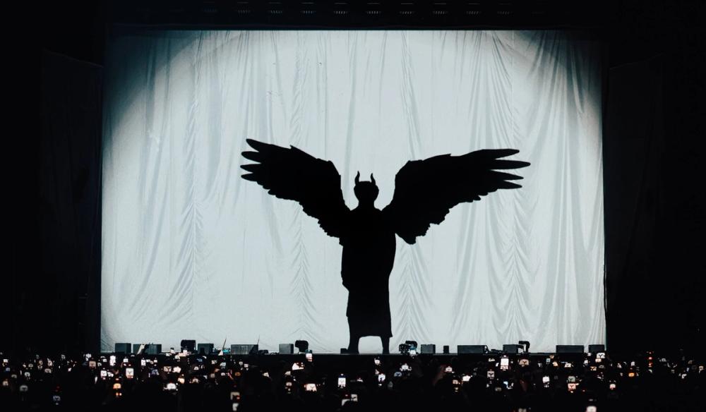 An audience stands facing a stage with their phones out recording. On the stage is a white kabuki drape with a silhouette of YUNGBLUD on it with horns and wings.