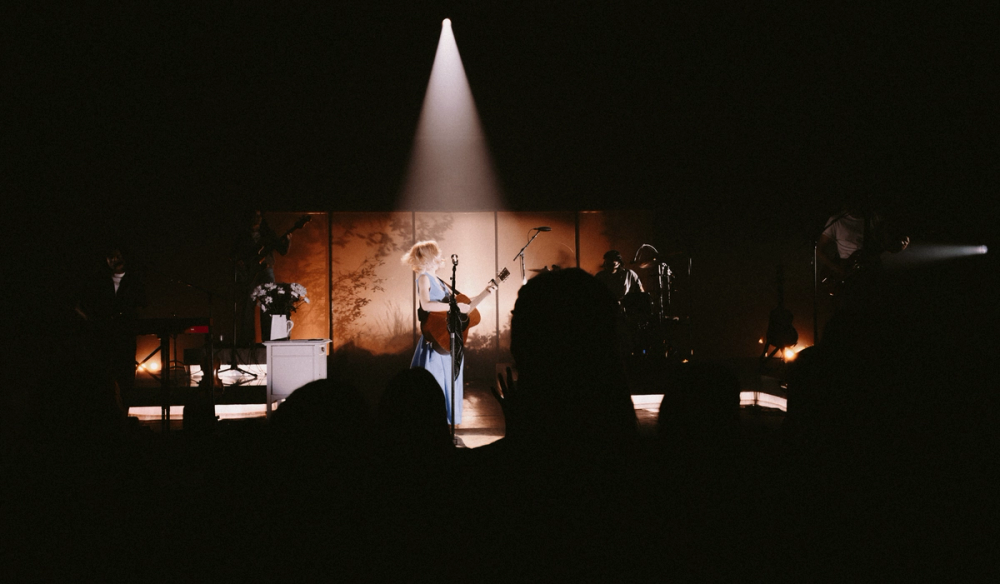 Maisie Peters spins around, center stage with her guitar in a blue dress. Behind her is an illuminated backdrop with shadows of plants.