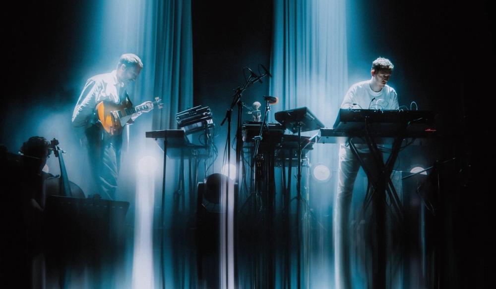 The two members of Maribou State stand on either side of the image with two blue light beams behind them