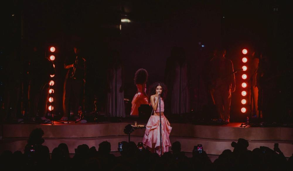 Olivia Dean stands on stage with a microphone, wearing a pink dress. Behind her is a mirror and two lighting towers