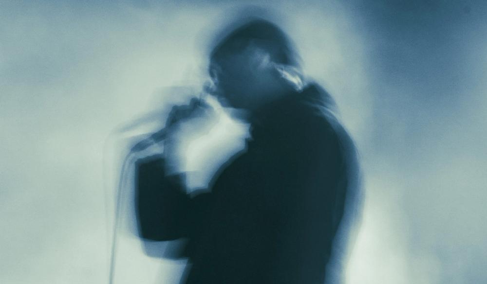 A monotone image of Corbin on stage holding a microphone, blue hues. Image is stylistically blurred