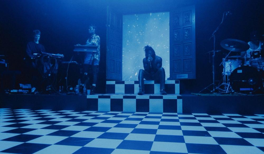 Obongjayar sits on the top of two stairs with a black and white checkerboard floor. To his left are right are bandmates including studio friend and talented musician Marla Kether. Behind Obongjayar is a set of antique doors which are flung open to reveal a galaxy behind him.