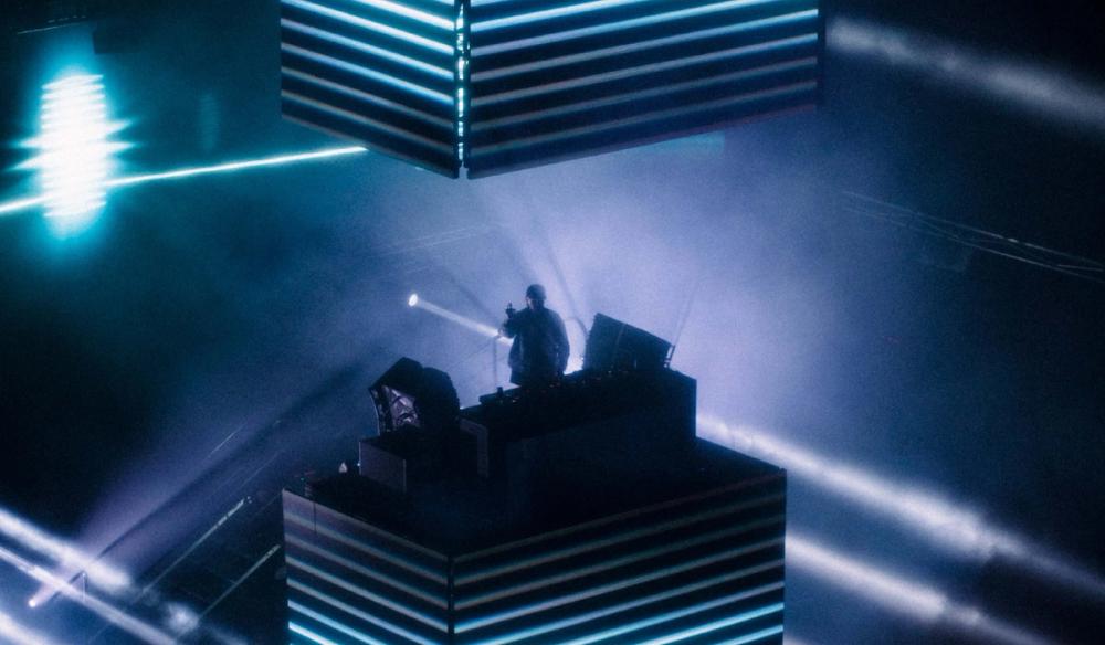 Dom Dolla Stands at a cube DJ riser with blue, glowing lines of light all around him.