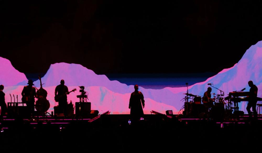 Alicia Keys stands centrally on stage. Her and her band are silhouetted by a large screen behind them that expands beyond the image, showing a horizon-line of purple and pink mountains.