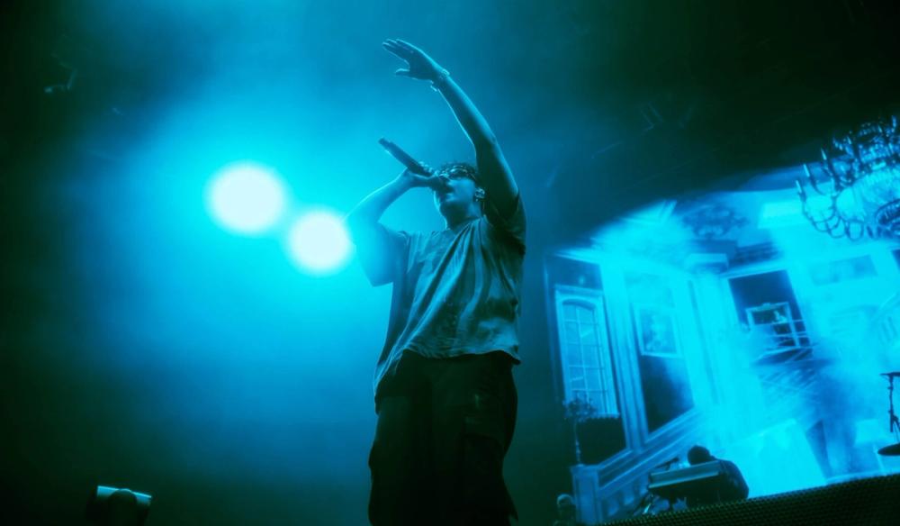 Artemas stands in blue lighting singing with a screen behind him with a mansion-like house on it.