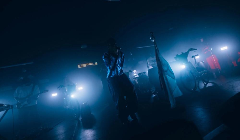 Jim Legxacy jumps on stage in blue lighting, bikes and a bus stop sign are around him as set