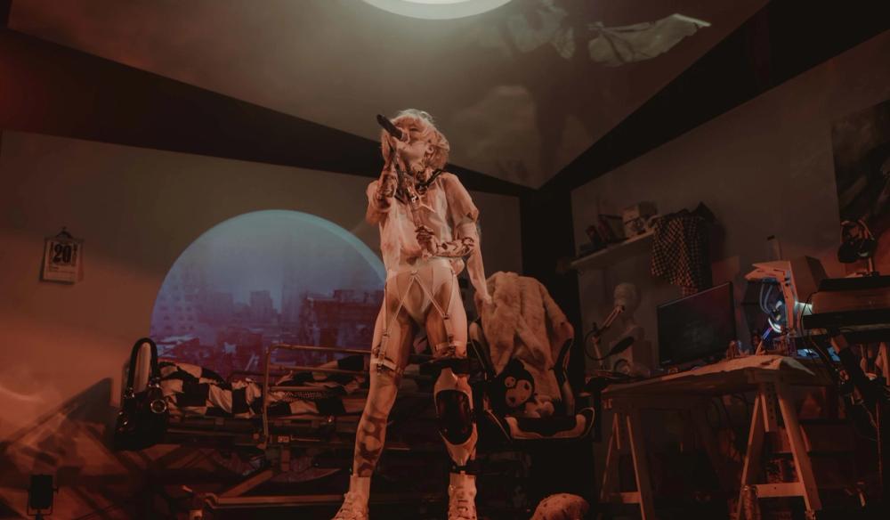 Artist Yeule stands center-frame holding a microphone. They are performing in front of a hospital style bed with a circle projection of a cityscape behind it. To the right side of the photo is a desk with glowing computer objects.