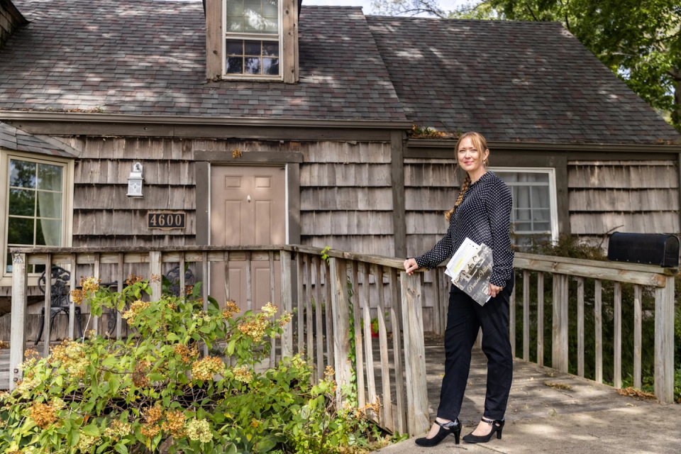 A woman stands at the wooden fence of a shingle-clad house numbered 4600, holding a folded newspaper.