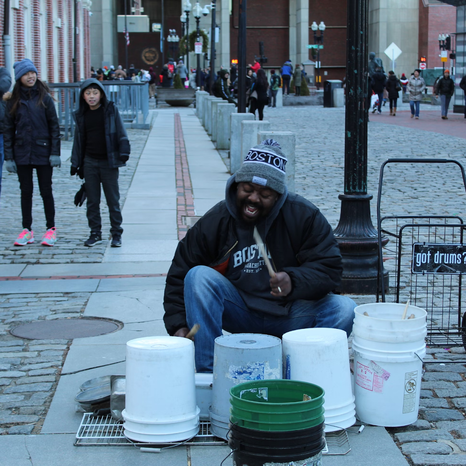 A street drummer plays on upturned plastic buckets on a cobbled city street while passers-by watch.