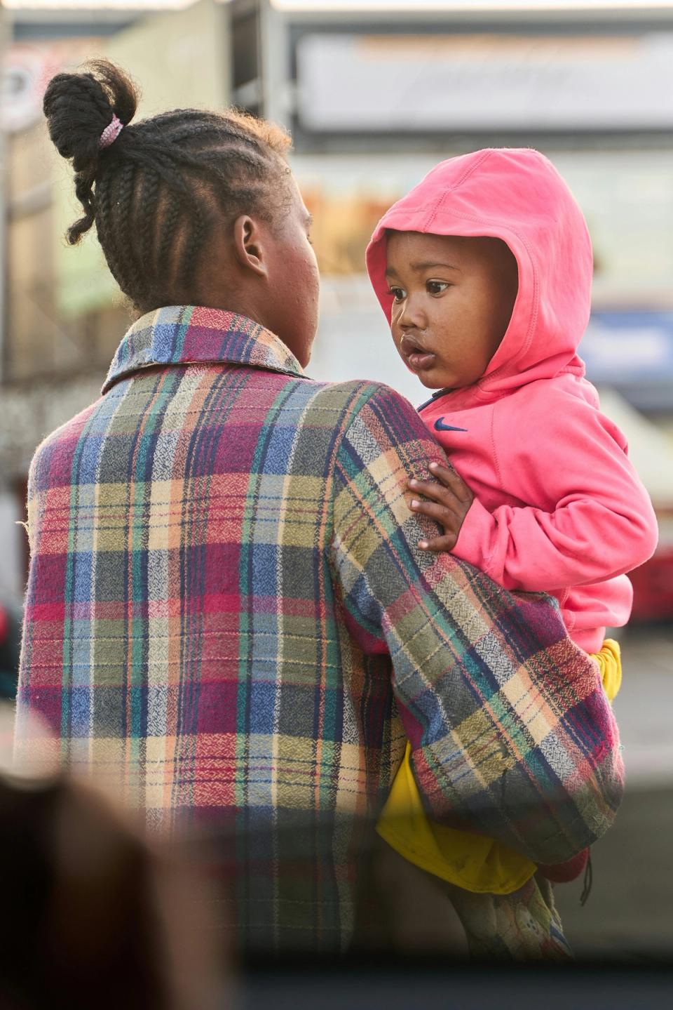 A woman in a plaid coat, seen from behind, carries a young child in a pink hooded top who looks back over her shoulder.