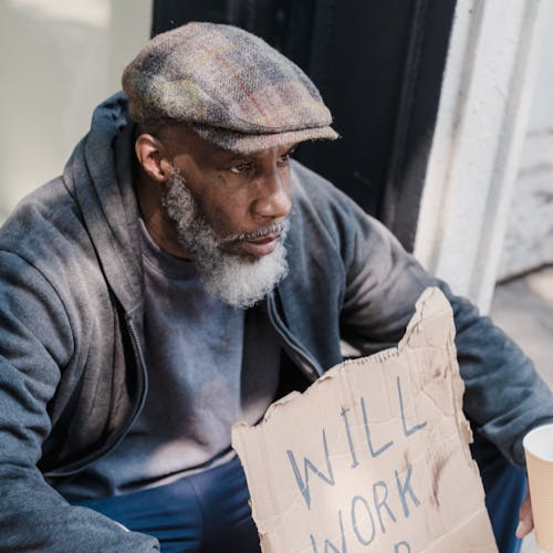 A man in a flat cap and hooded jacket sits on a pavement holding a torn cardboard sign that reads “WILL WORK”.