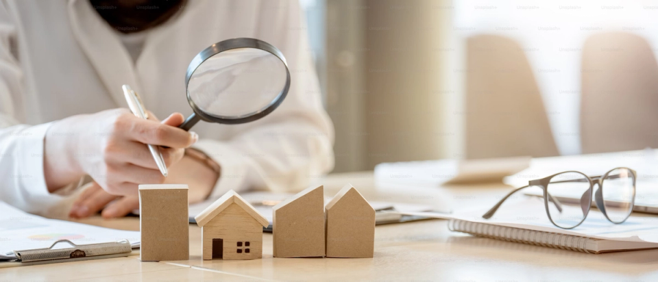 A row of small wooden model houses on a desk, with a person behind them holding a magnifying glass and a pen.