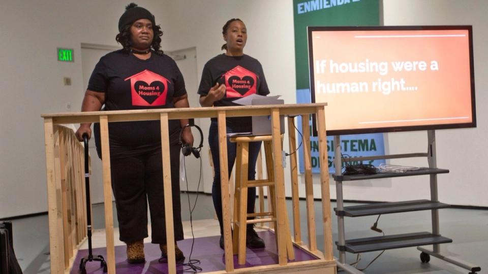 Two women in black Moms 4 Housing T-shirts stand inside a wooden frame on a gallery stage; one speaks while a screen beside them reads “If housing were a human right…”.