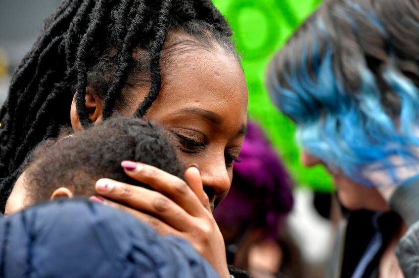 A woman with long locs rests her face against the head of the child she is holding, eyes lowered, at a crowded outdoor gathering.