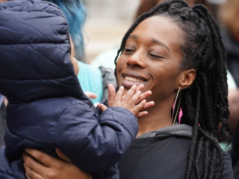 Dominique Walker, smiling, with long locs and hoop earrings holding a toddler in a navy puffer jacket, who reaches up to touch her face; a crowd is blurred behind them.