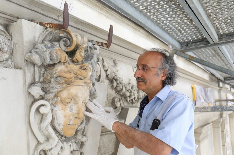 A conservator wearing gloves and a light blue shirt carefully restores an ornate sculptural detail featuring a carved face on a historic building, working from a scaffold.