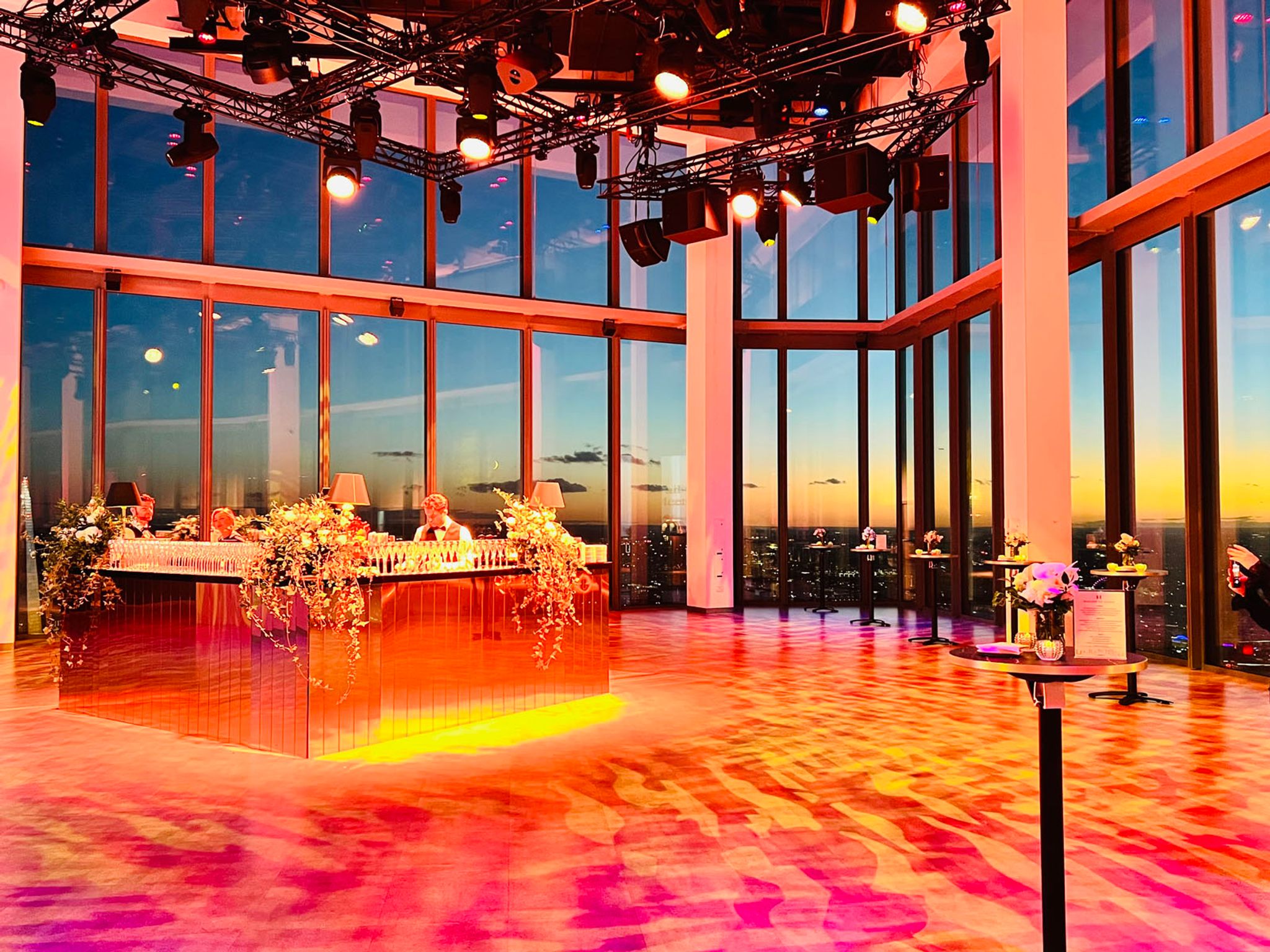 The Horizon 22 event space bathed in redish light at twilight. A golden square panelled bar is set up in the middle of the room and small round cocktail tables with flowers on them are evenly dotted around the space.