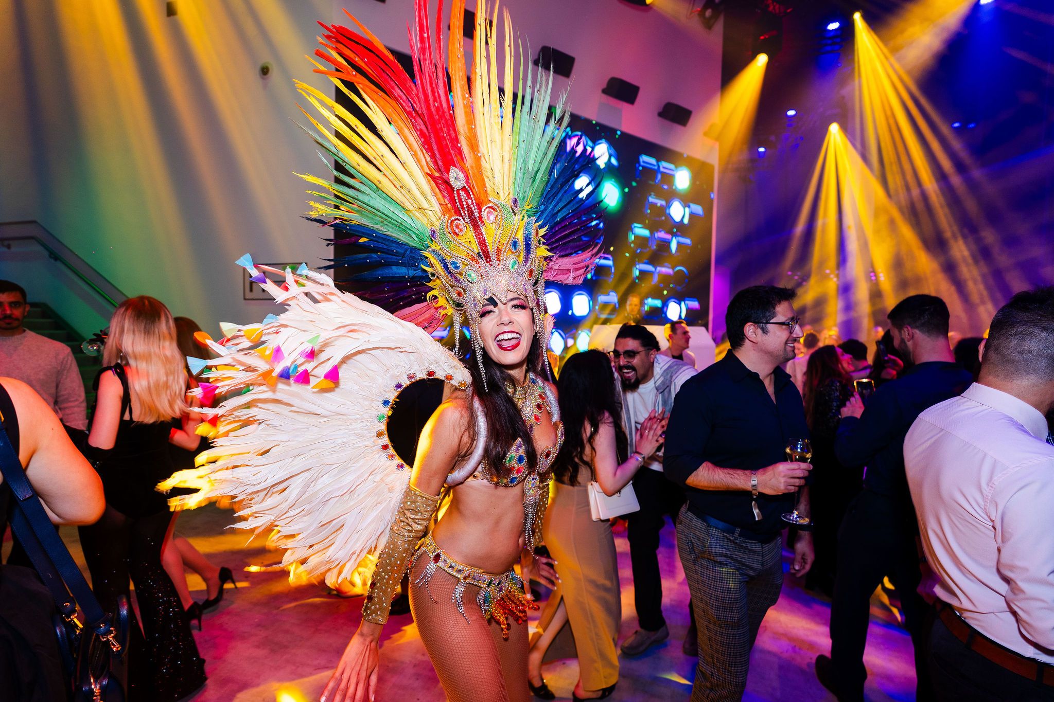 Smiling dancer in a bejewelled carnival style costume with a large rainbow feathered headdress posing among party guests on a lit dance floor.