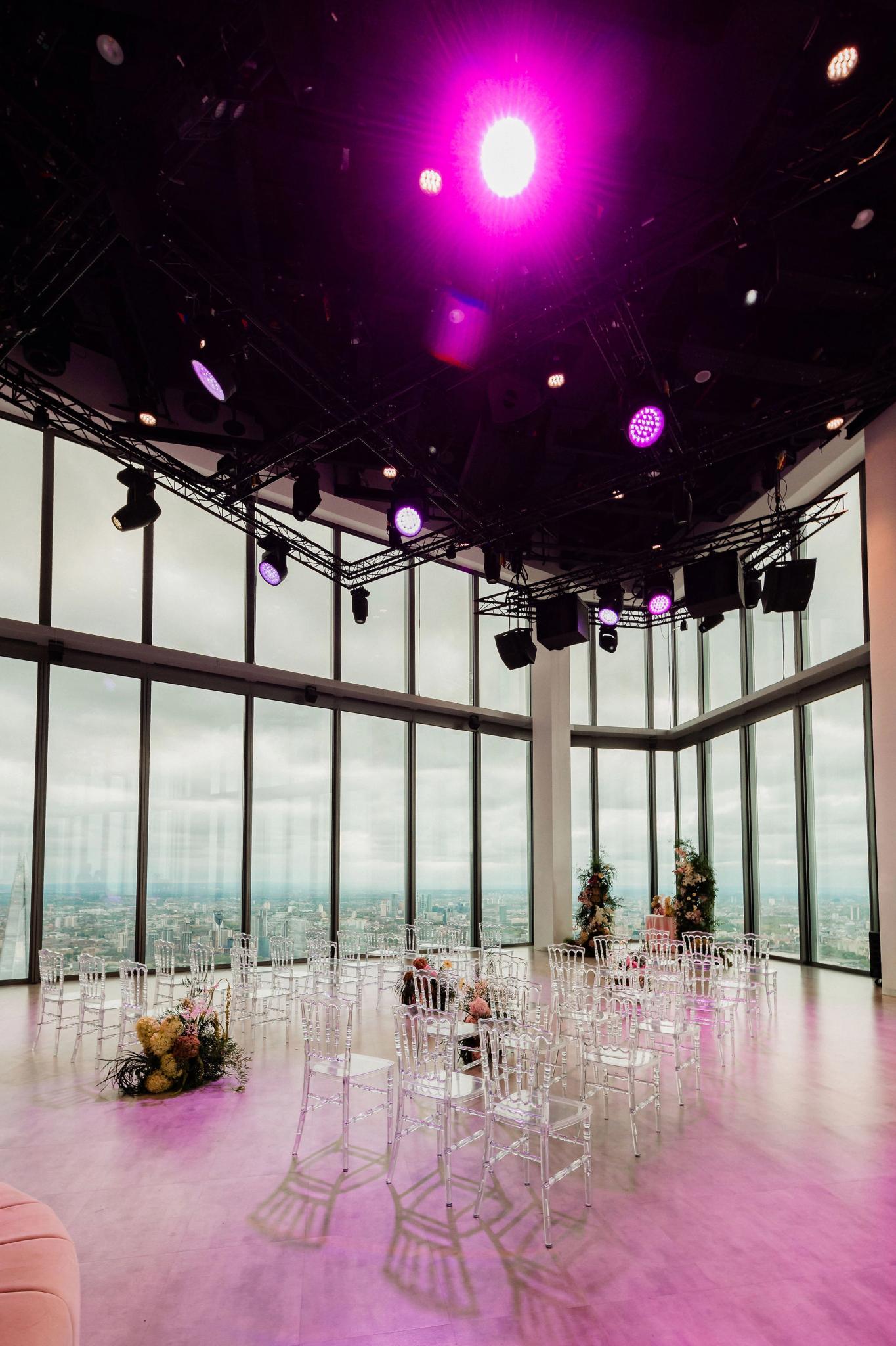 An indoor wedding ceremony setup with transparent chairs arranged in rows, surrounded by floor-to-ceiling windows showing a city skyline, and purple stage lighting illuminating the space from above.