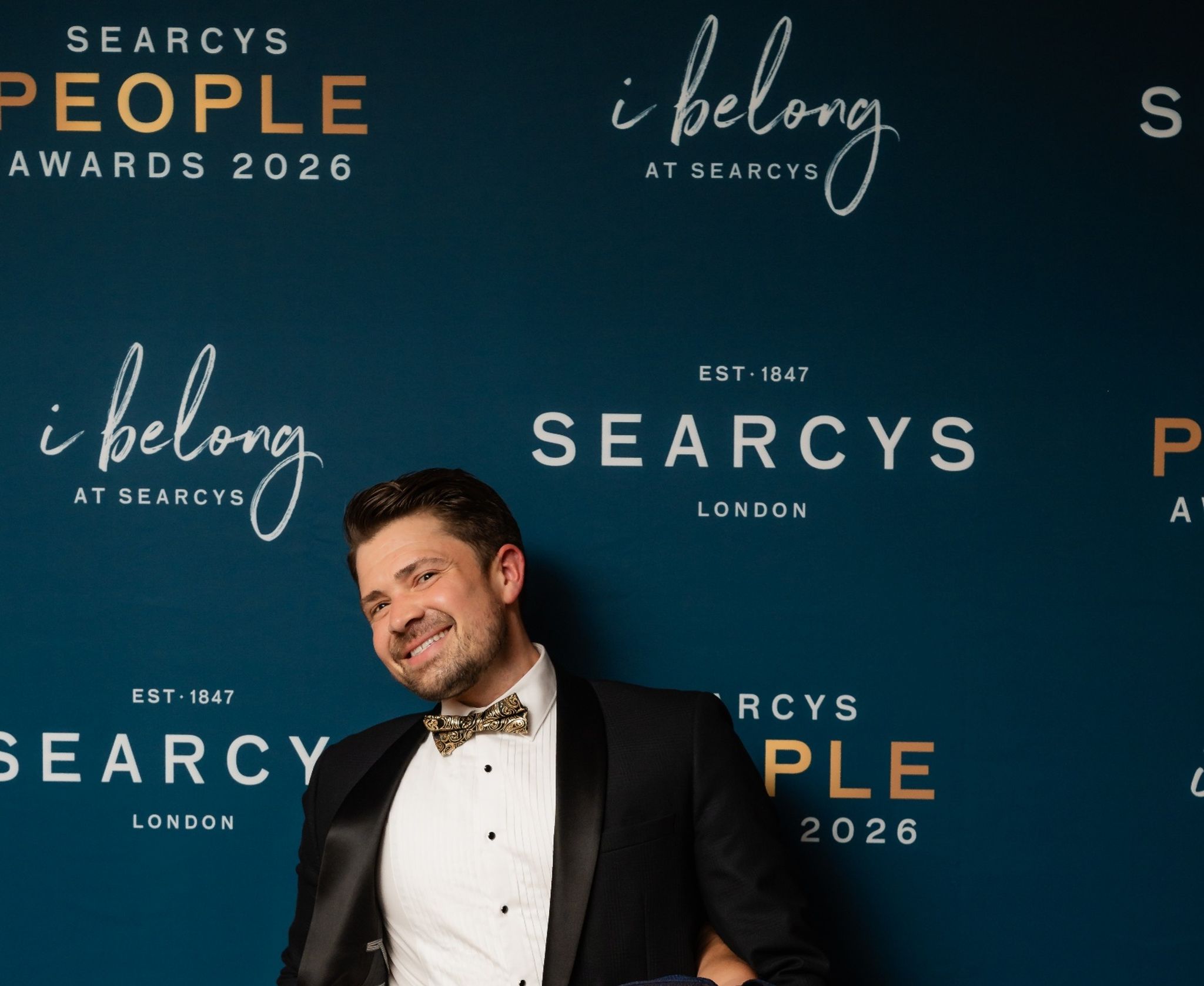 A man in a tuxedo in front of a Searcy's hoarding