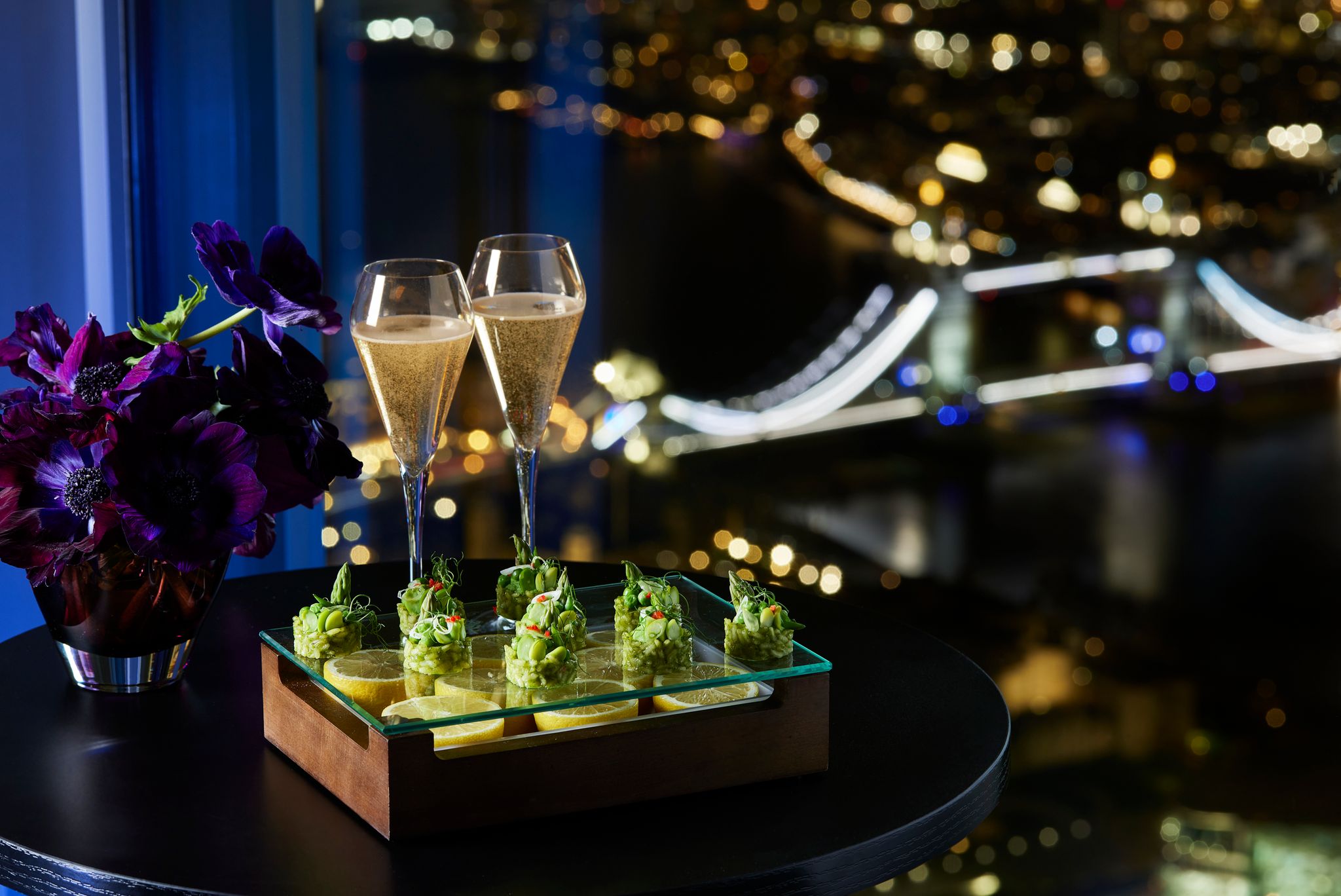 Bride and groom share a kiss during their wedding ceremony in a glass-walled venue high above the city, surrounded by guests and floral arrangements.