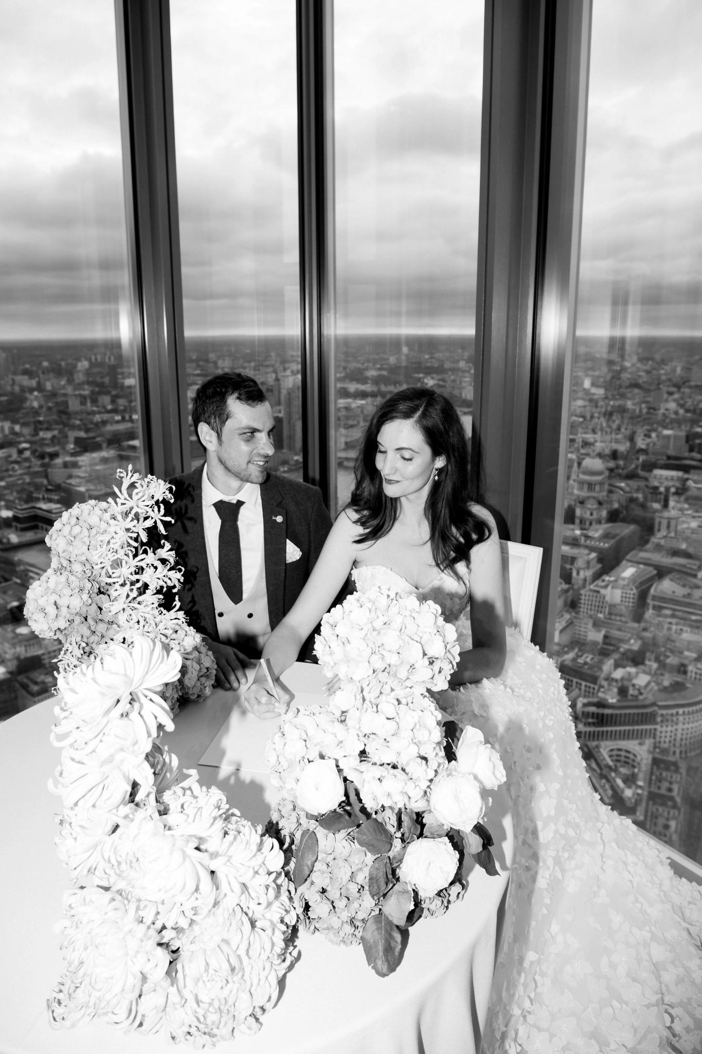 A black and white photo of a bride and groom sitting together at a table adorned with large floral arrangements, with floor-to-ceiling windows overlooking a cityscape in the background.