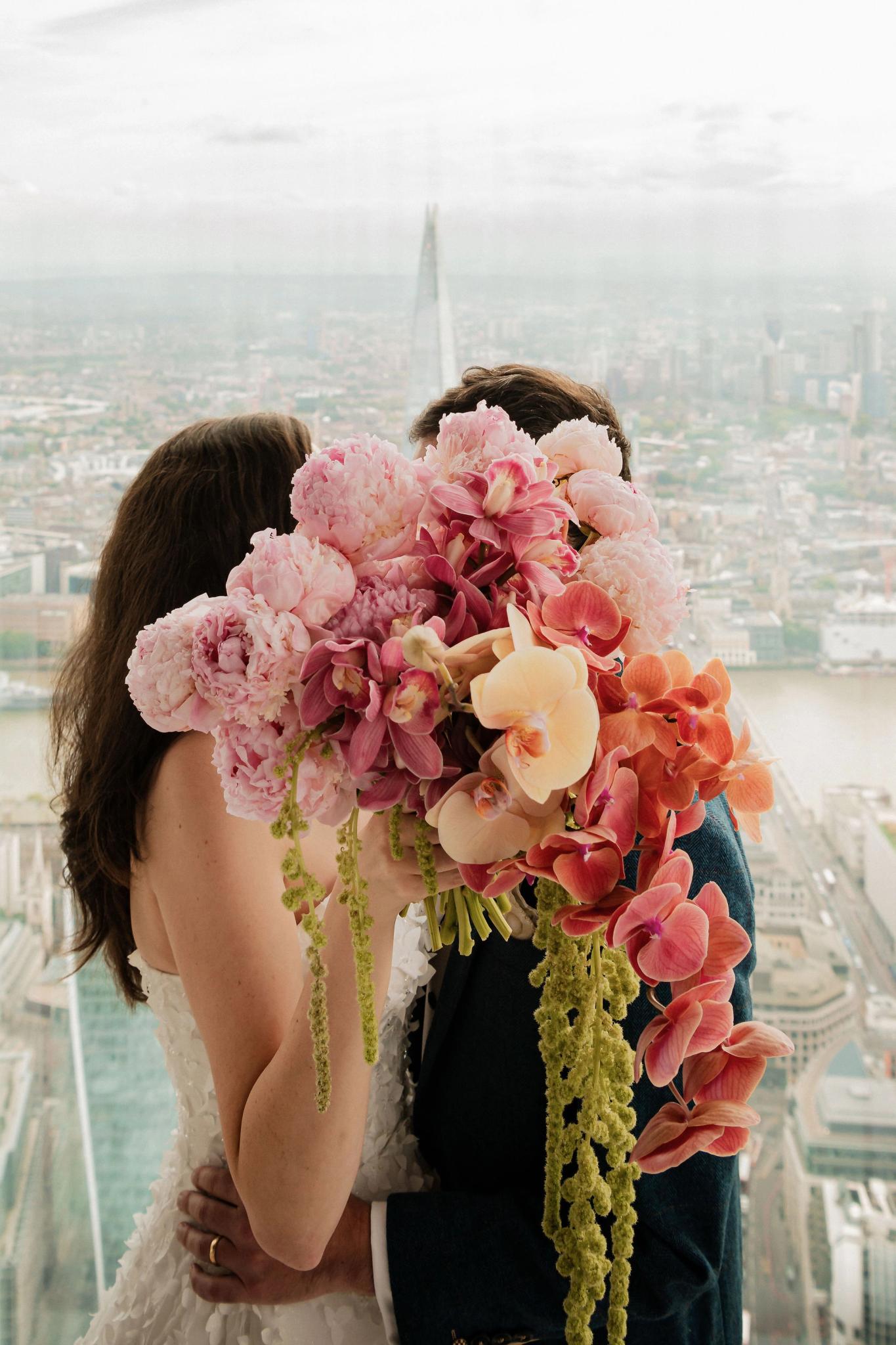 A couple embracing in wedding attire, standing in front of a city skyline view while holding a large bouquet of pink and peach flowers that obscures their faces.