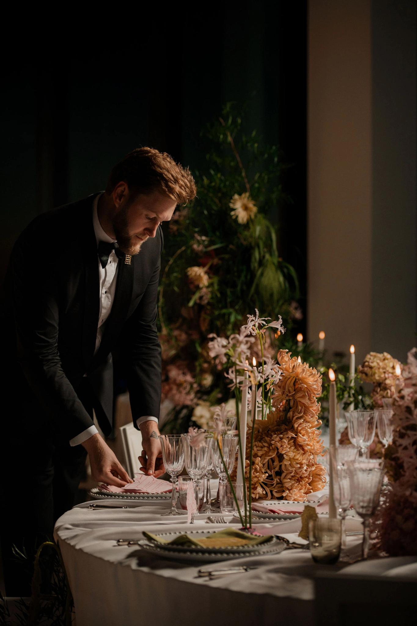 A man dressed in a black tuxedo is carefully arranging table settings at an elegantly decorated event, with floral centerpieces and candles on a round table in a dimly lit room.