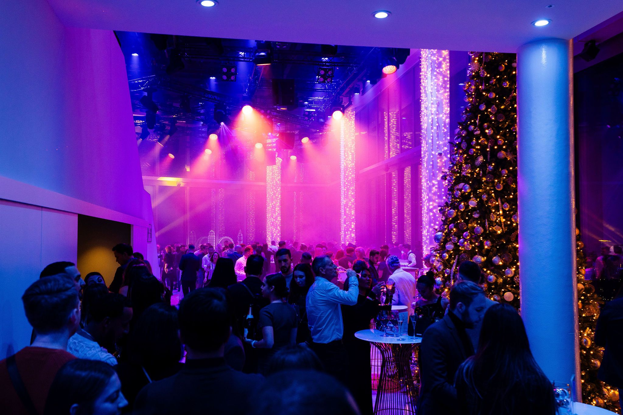 Crowded indoor New Year’s Eve party with purple and blue stage lighting, people mingling around tall tables, and a large decorated Christmas tree on the right.