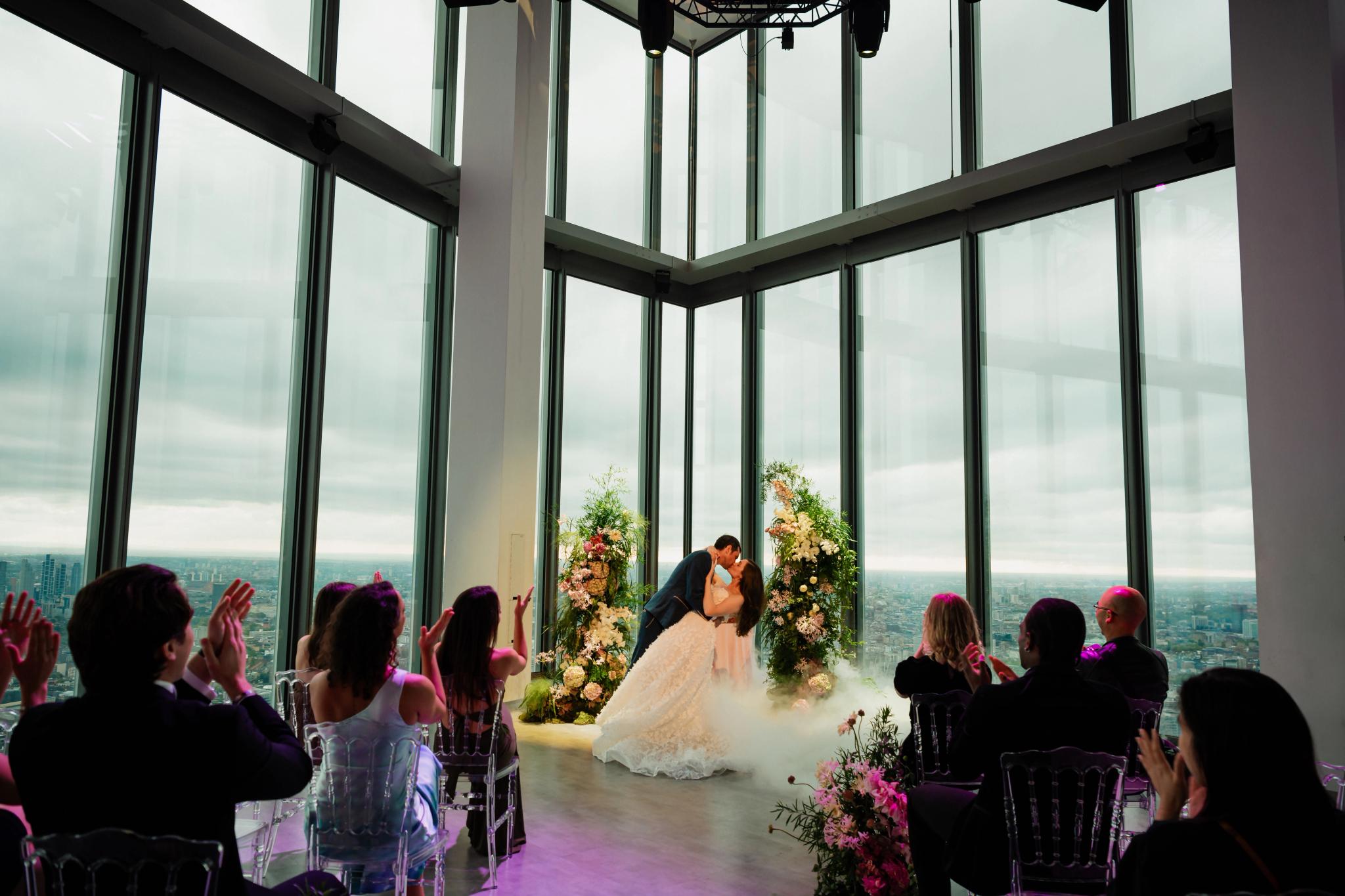 Bride and groom share a kiss during their wedding ceremony in a glass-walled venue high above the city, surrounded by guests and floral arrangements.