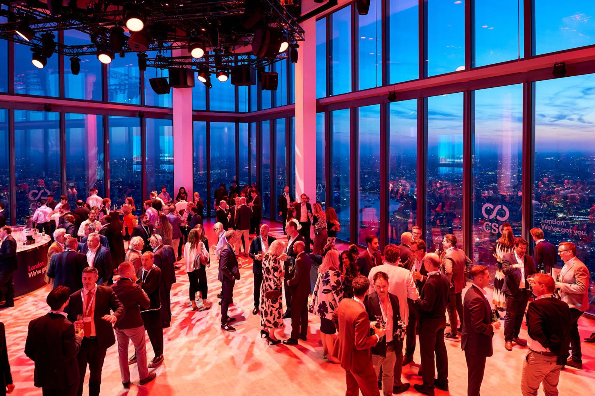 A large group of people seen mingling for an event in the venue space at Horizon 22 at twilight. The room is lit with lights that give off a redish tinge. 