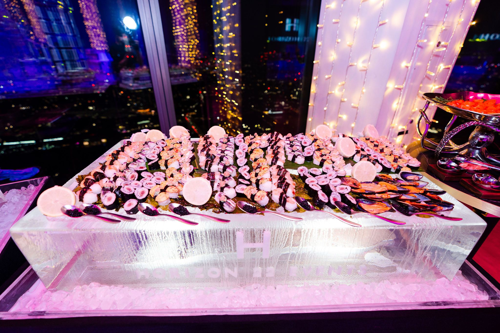 Buffet display of sushi and seafood arranged on a large rectangular ice block, with lemon slices and decorative lights reflected in the window behind.