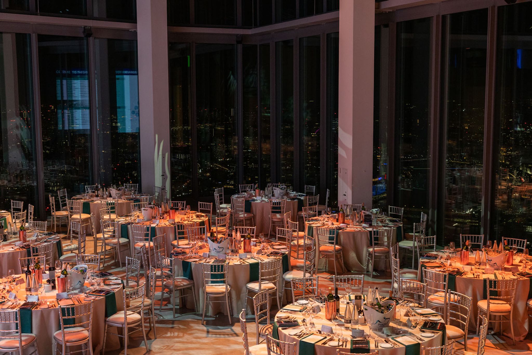 The Horizon 22 events hall at night with 9 tables laid for a fine dining experience, with a beige tablecloth. On the tables are wine glasses, knifes and folks and bottles of champagne. 