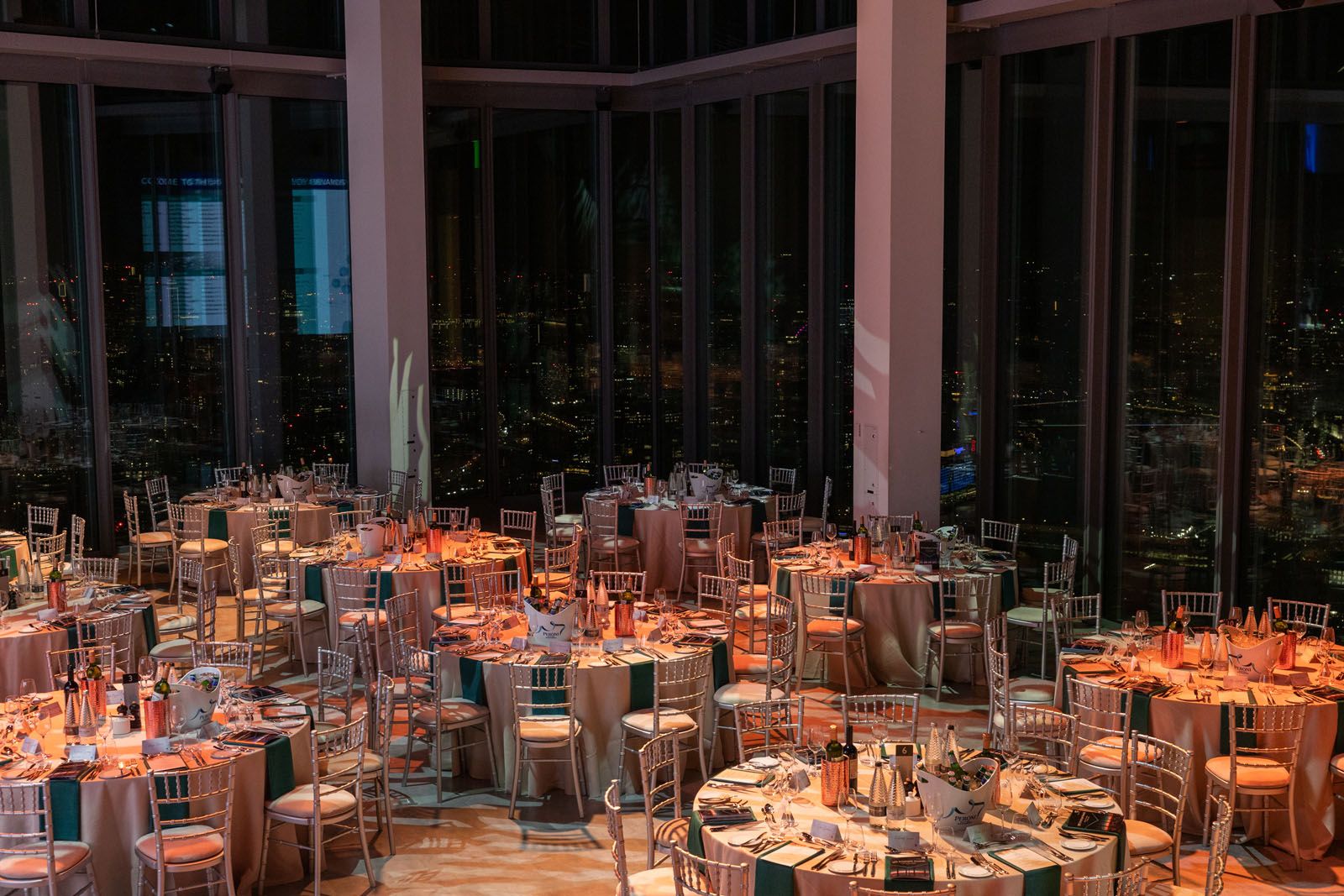 The Horizon 22 events hall at night with 9 tables laid for a fine dining experience, with a beige tablecloth. On the tables are wine glasses, knifes and folks and bottles of champagne.
