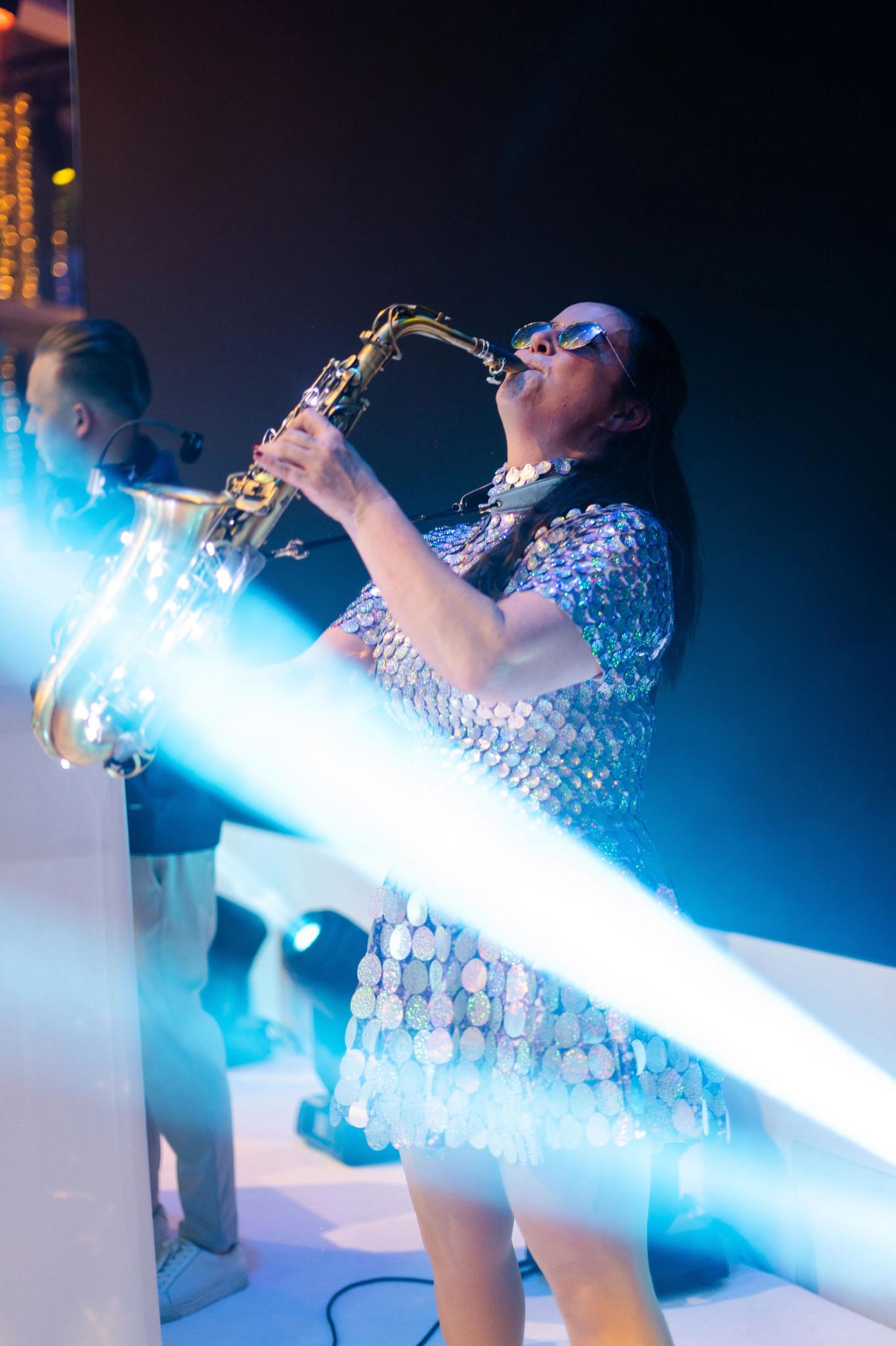 Female musician in a reflective sequin dress playing a saxophone on stage under bright blue and white beams of light.