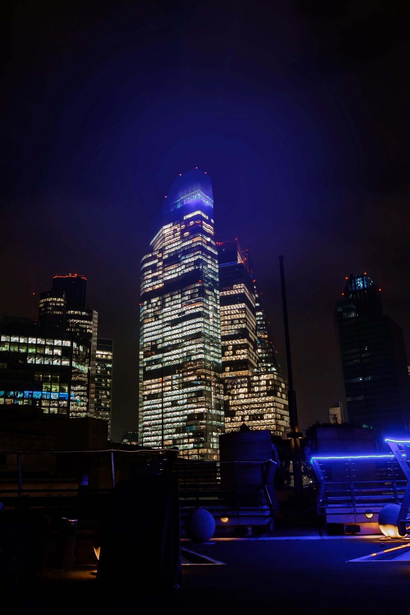 Close-up of a modern glass skyscraper at night, its many office windows brightly lit, rising into a dark sky with a soft bluish halo near the top
